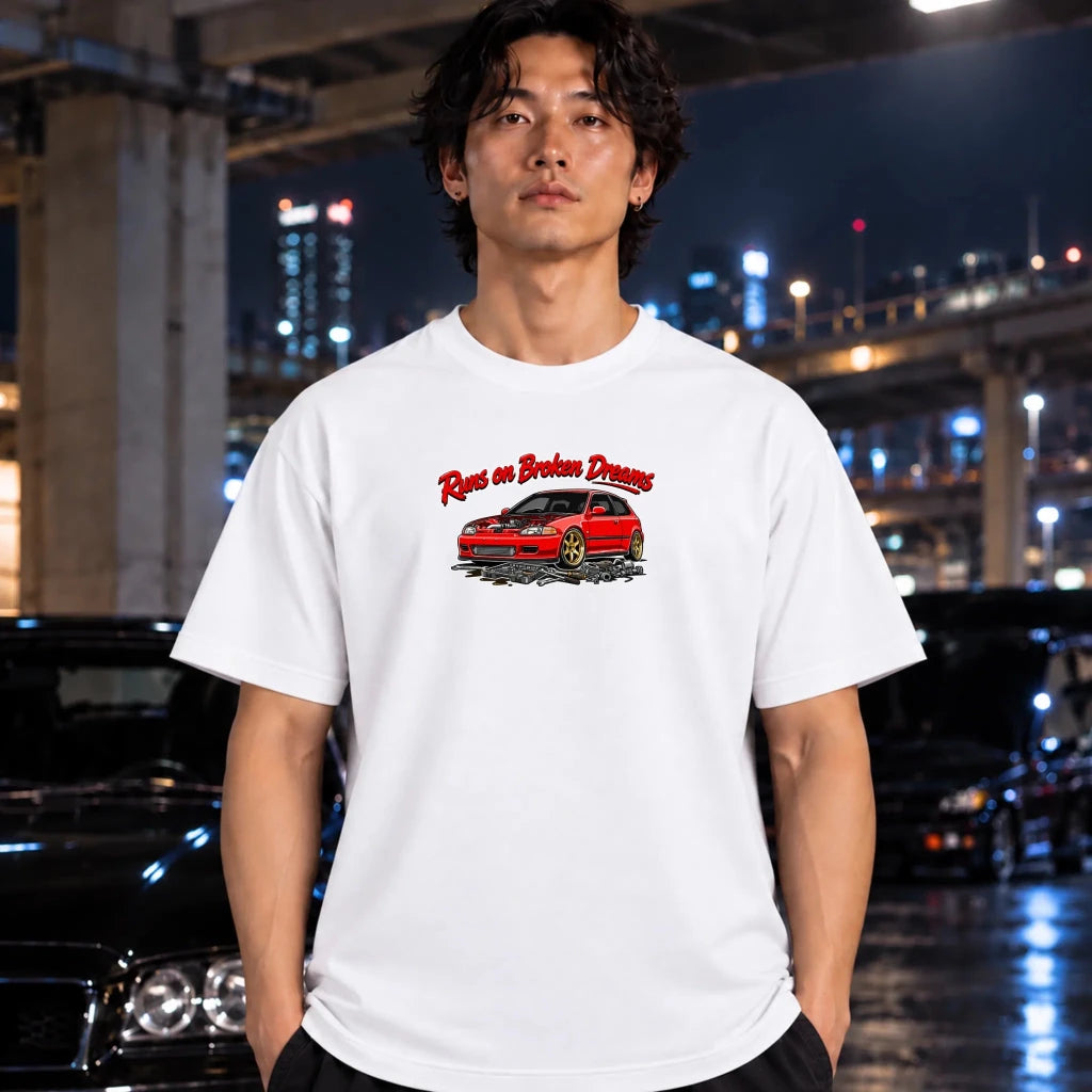 A man wearing a white graphic t-shirt with a red car and 'Runs on Broken Dreams' text, standing in a city environment at night with blurred cars and lights in the background.