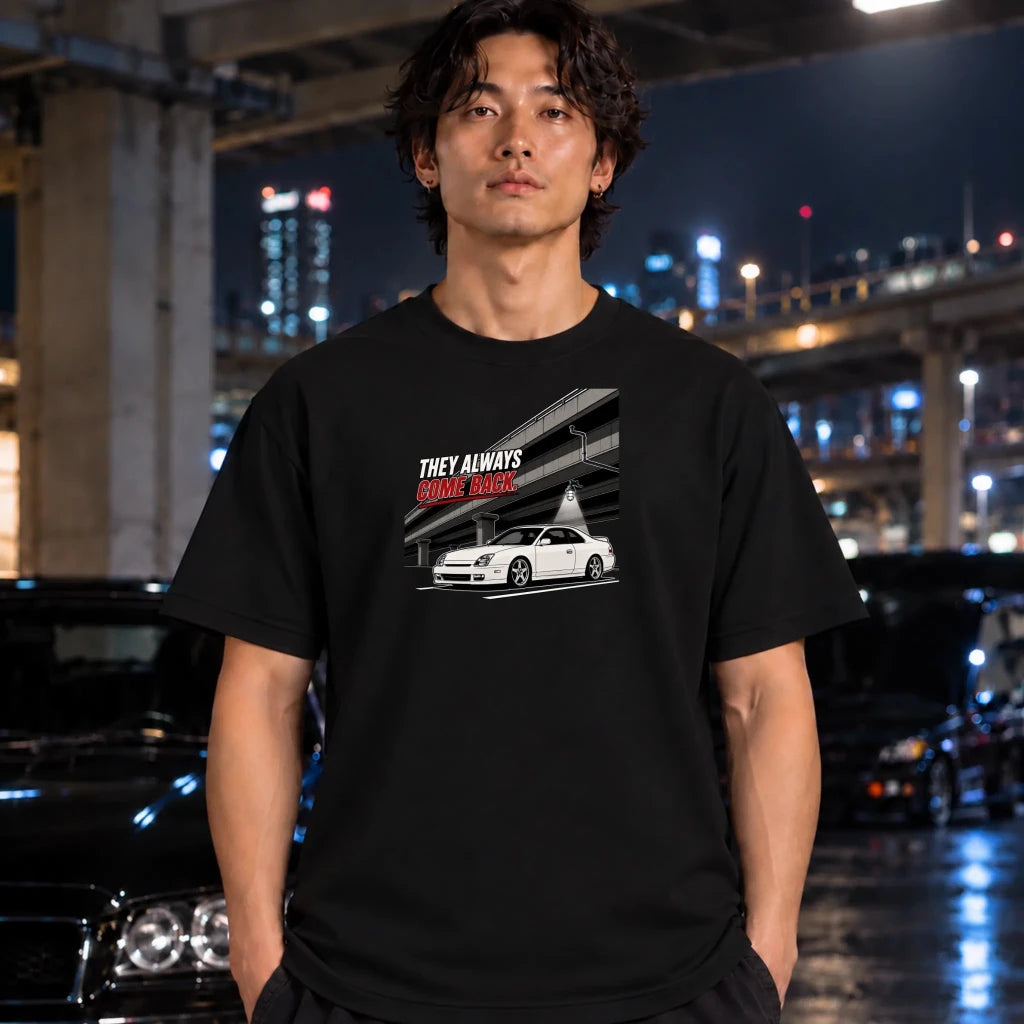 A man wearing a black graphic t-shirt with a white and red print of a car and the text 'THEY ALWAYS COME BACK' stands in a nighttime urban setting with blurred cars and city lights in the background.