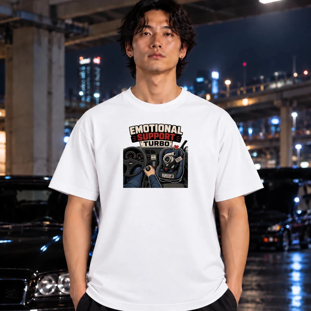 A man wearing a white graphic t-shirt with a 'EMOTIONAL SUPPORT TURBO' car dashboard print, standing in a wet, nighttime urban environment with cars and city lights in the background.