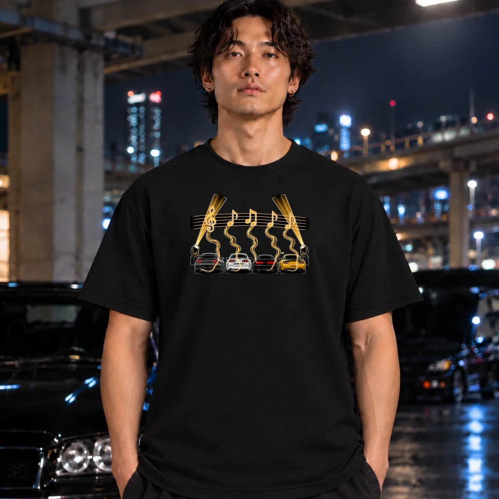 A man wearing a black graphic t-shirt with a yellow and white car design, standing in a nighttime urban setting with blurred cars and city lights in the background.