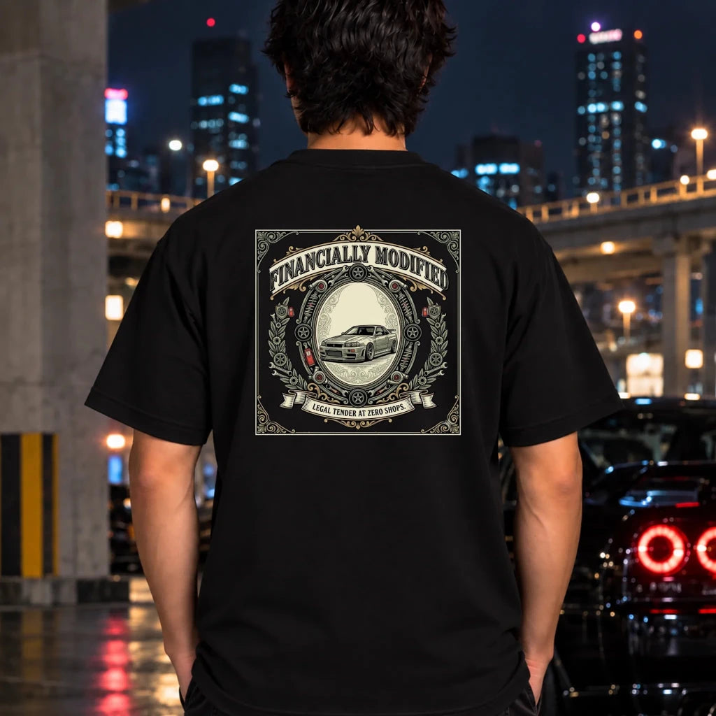 A man viewed from behind wearing a black t-shirt with a large vintage-style graphic on the back that reads 'FINANCALLY MODIFIED' and features a car illustration. The background is a blurred nighttime city street scene with lights and a car taillight.