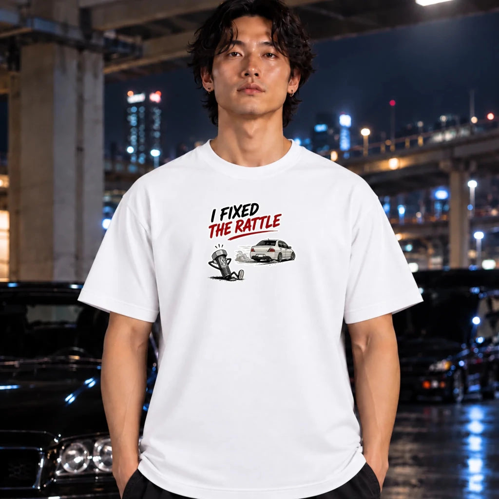 A man wearing a white graphic t-shirt with the text 'I FIXED THE RATTLE' and a small car/demon illustration, standing in a city environment at night with cars and lights in the background.