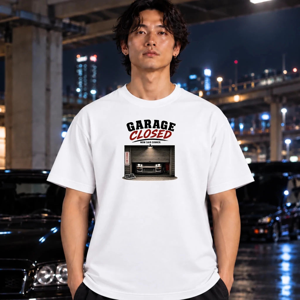 A man wearing a white  short-sleeve graphic t-shirt. The graphic features the text 'GARAGE CLOSED' in black and red, above an illustration of a car parked inside a garage.