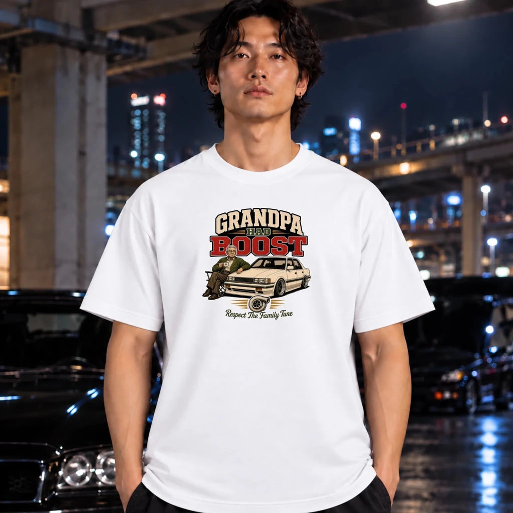 A man wearing a white graphic t-shirt with a 'grandpa had boost' car illustration, standing in a city environment at night with blurred cars and lights in the background.