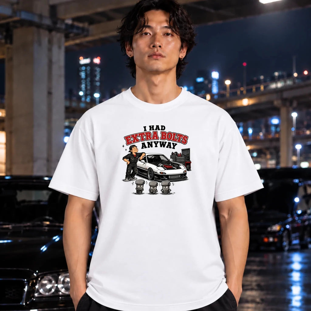 A man wearing a white graphic t-shirt with a cartoon-style print of a car, a character, and the text 'I HAD EXTRA BOLTS ANYWAY' stands in front of a blurred night city background with cars.