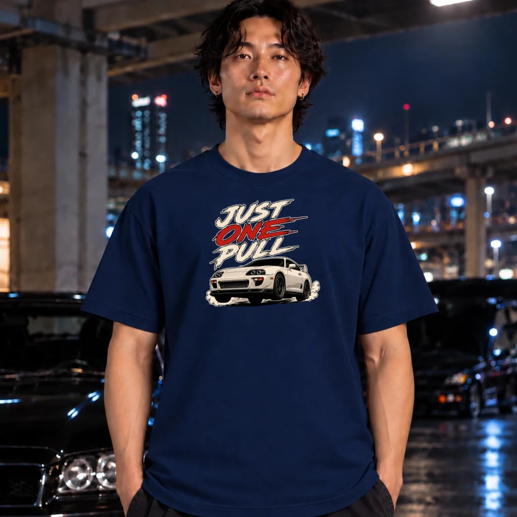 A man wearing a navy blue graphic t-shirt with a white car and 'JUST ONE PULL' text, standing in a wet urban night scene with blurred cars and city lights in the background.