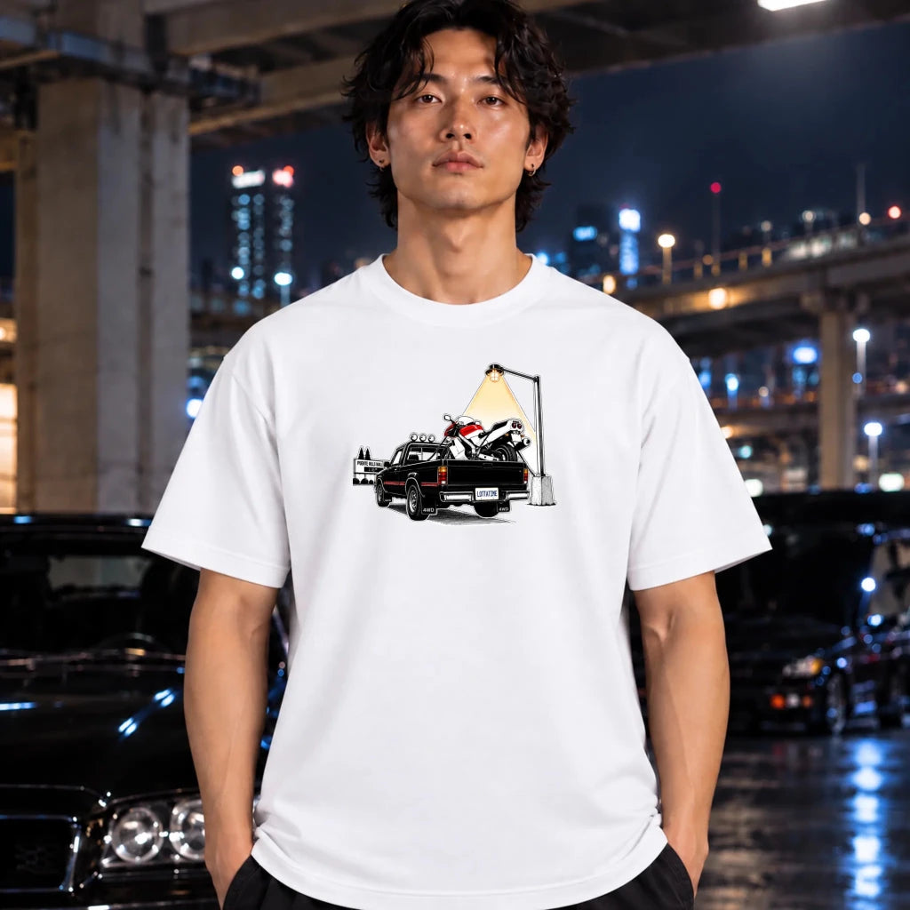 A man wearing a white crewneck t-shirt with a graphic print of a black pickup truck carrying a motorcycle, standing in a city environment at night with blurred cars and lights in the background.