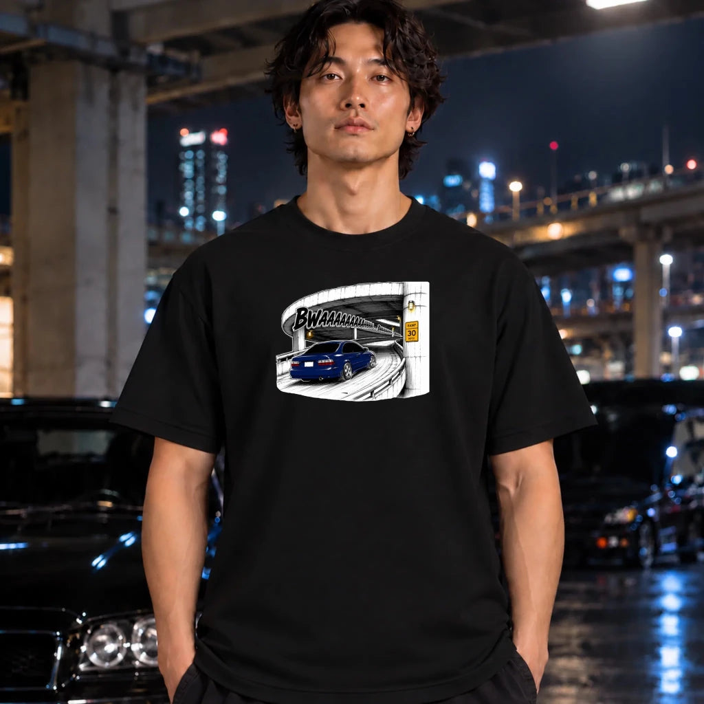 A man wearing a black graphic t-shirt featuring a blue sports car on a on ramp, standing in a nighttime urban setting with blurred cars and city lights in the background.