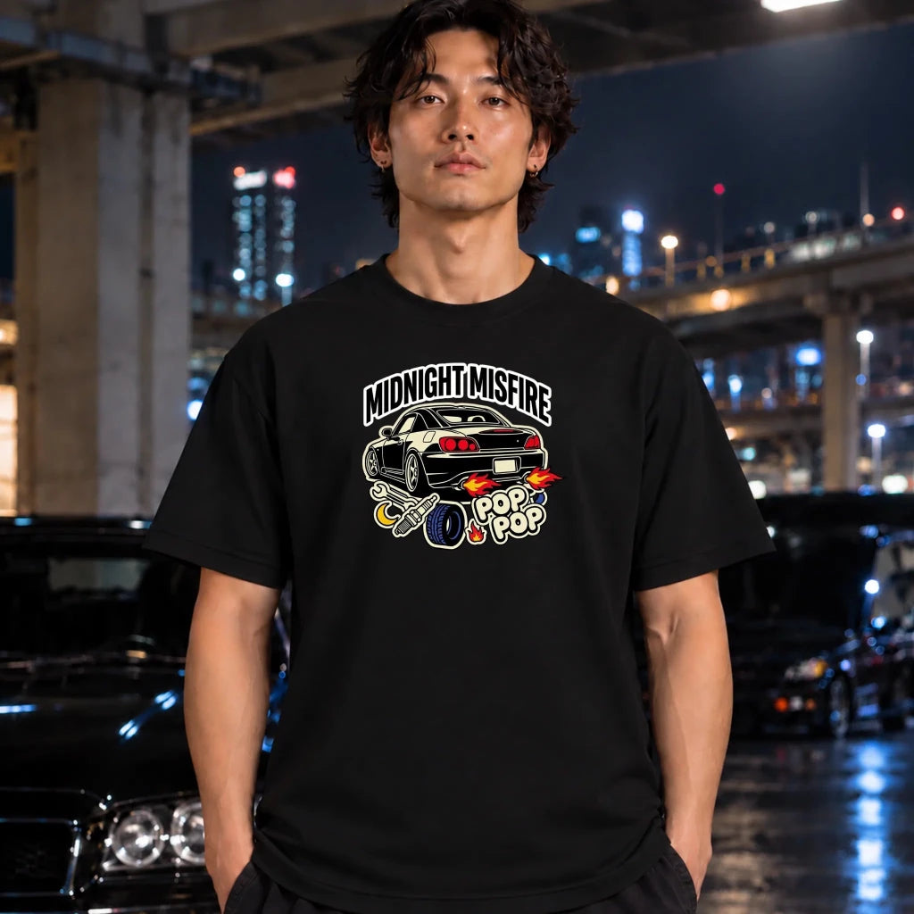 A man wearing a black graphic t-shirt with a 'Midnight Misfire' car design, standing in a wet urban night setting with blurred cars and city lights in the background.