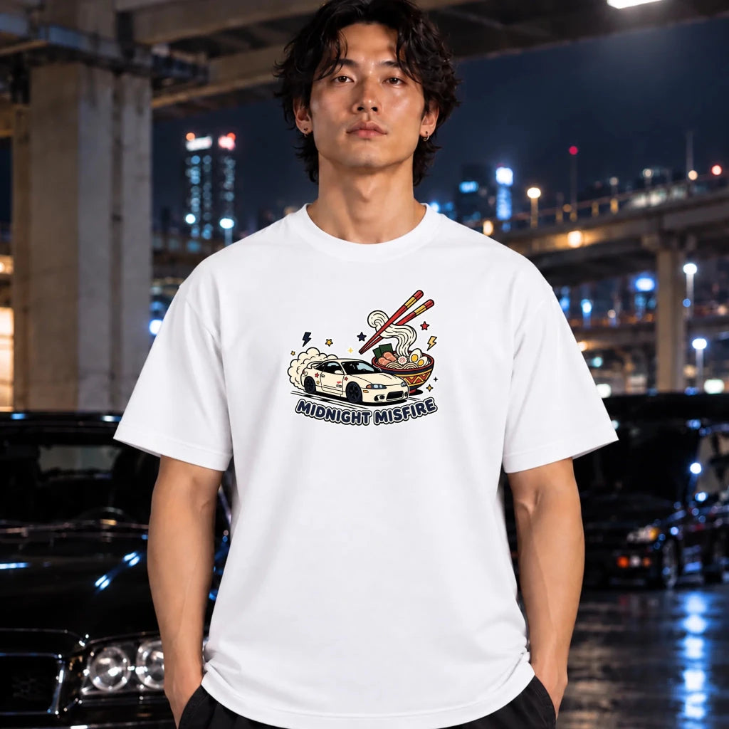 A man wearing a white graphic t-shirt with a 'Midnight Misfire' car illustration, standing in front of a blurred night city background with cars.