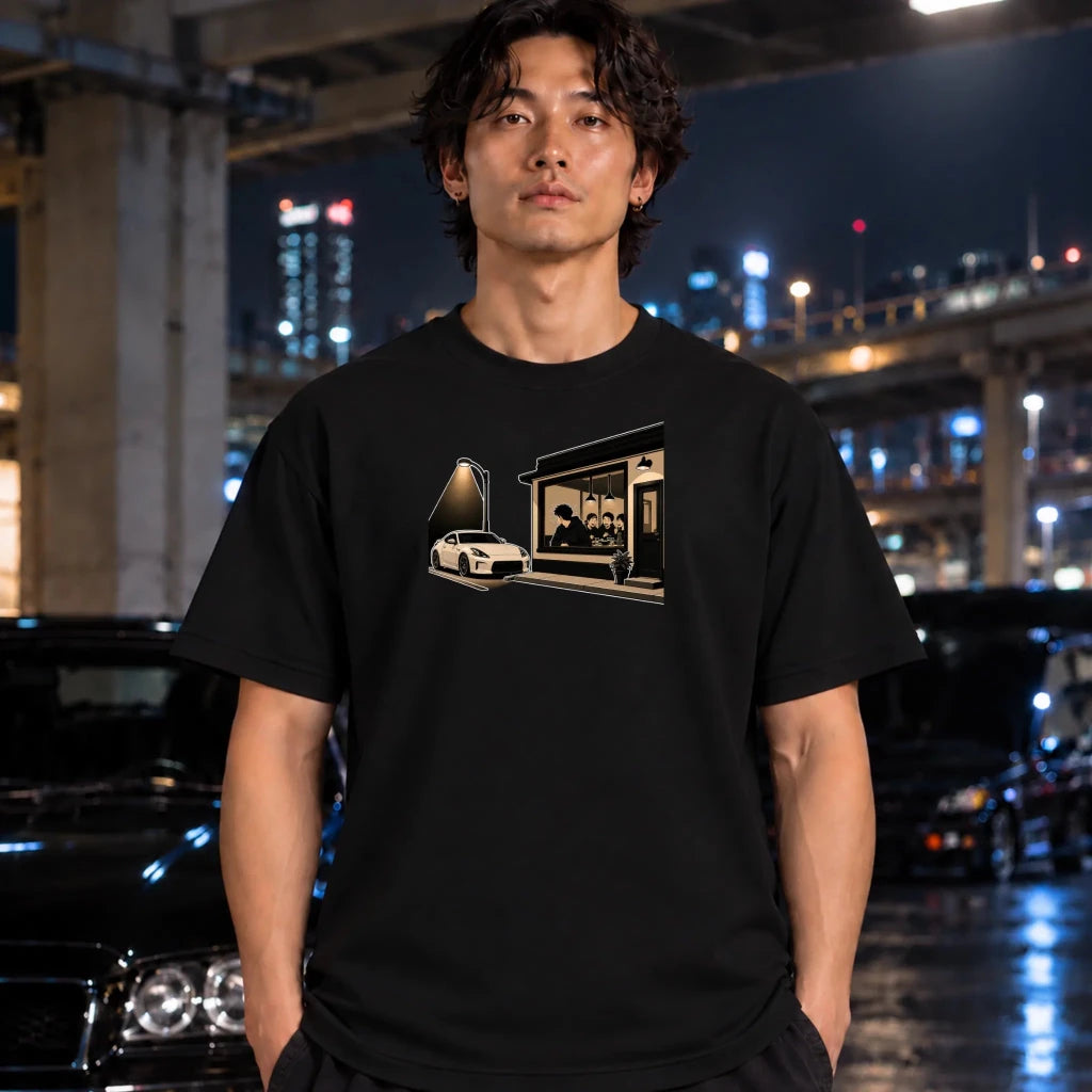 man wearing a black t-shirt, featuring a graphic print of a white sports car parked outside a dimly lit cafe. 