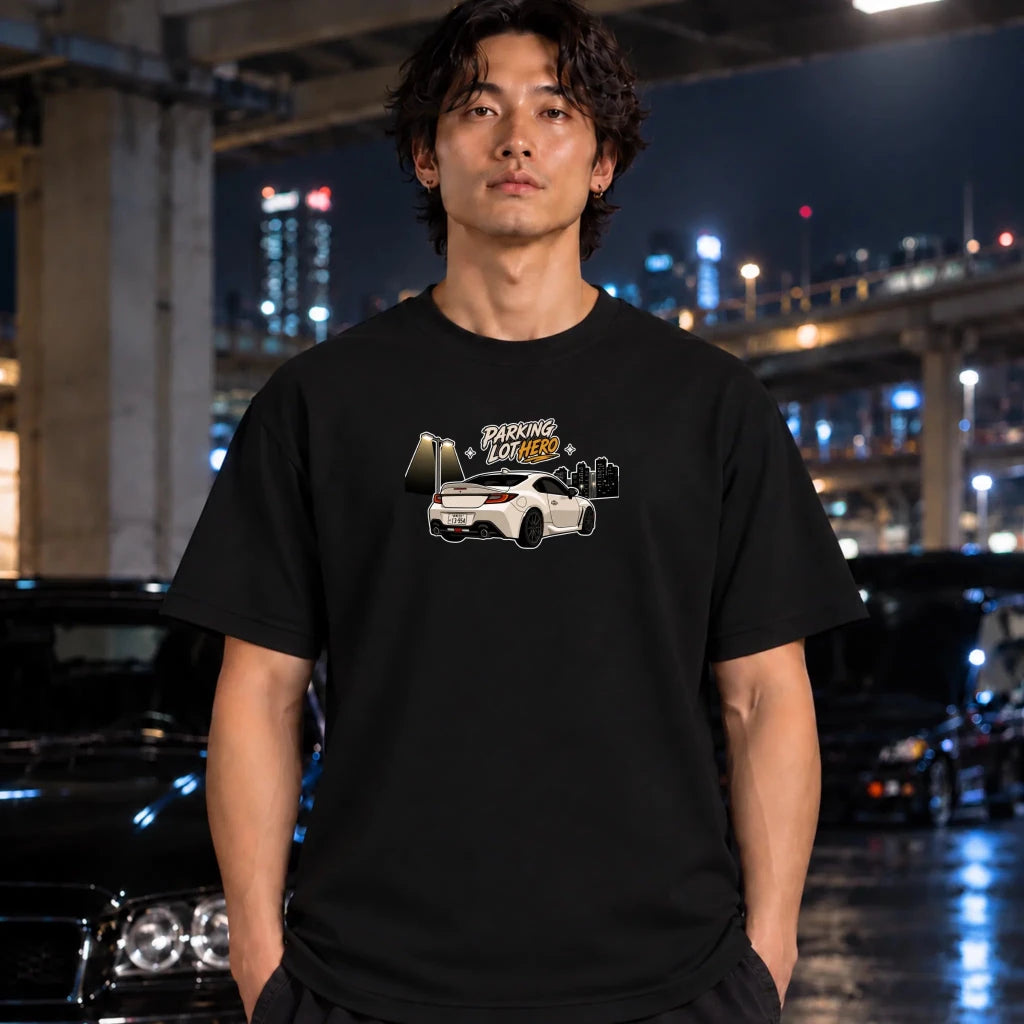 A man wearing a navy blue t-shirt with a graphic print of a white sports car and the text 'PARKING LOT HERO' stands in a city parking structure at night.