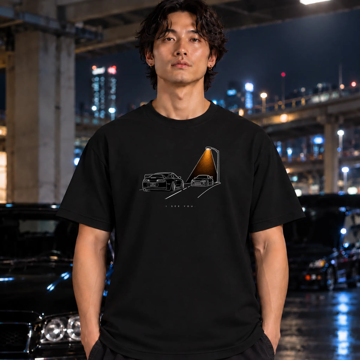 A man wearing a black graphic t-shirt with a minimalist car illustration, standing in a wet urban night scene with city lights and cars in the background.