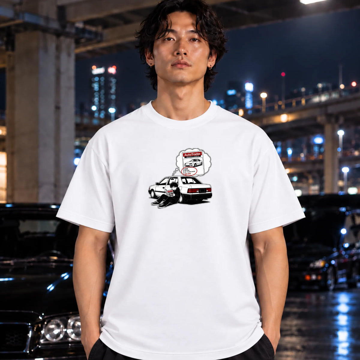 A man wearing a white graphic t-shirt featuring a cartoon sedan car, standing in front of a blurred night city background with cars.