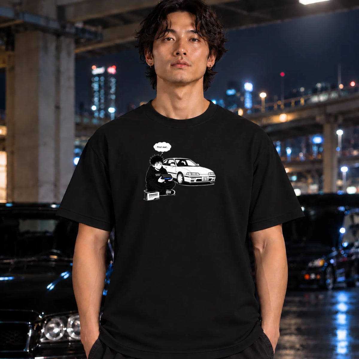 A man wearing a black graphic t-shirt with a white illustration of a person and a car, standing in a wet urban night setting with cars and city lights in the background.