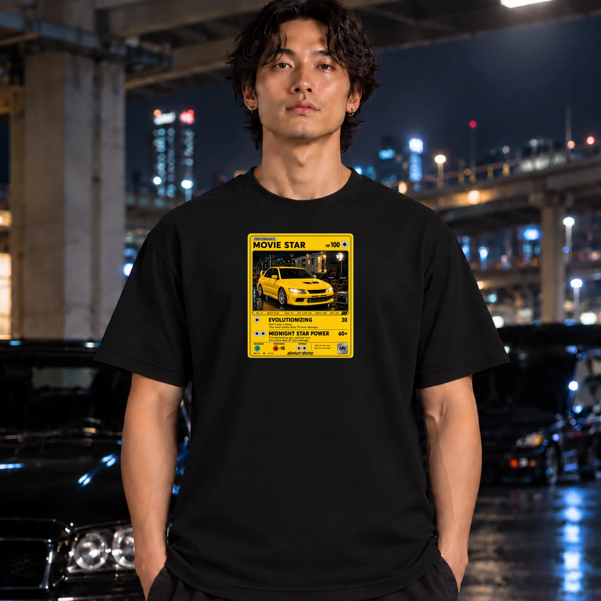 A man wearing a black t-shirt with a yellow graphic print that reads 'MOVIE STAR' and features a yellow car illustration, standing in a nighttime urban setting with blurred cars and city lights in the background.