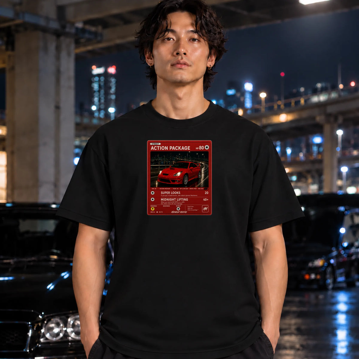 A man wearing a black t-shirt with a red and white graphic print on the chest, standing in a city environment at night with blurred cars and lights in the background.