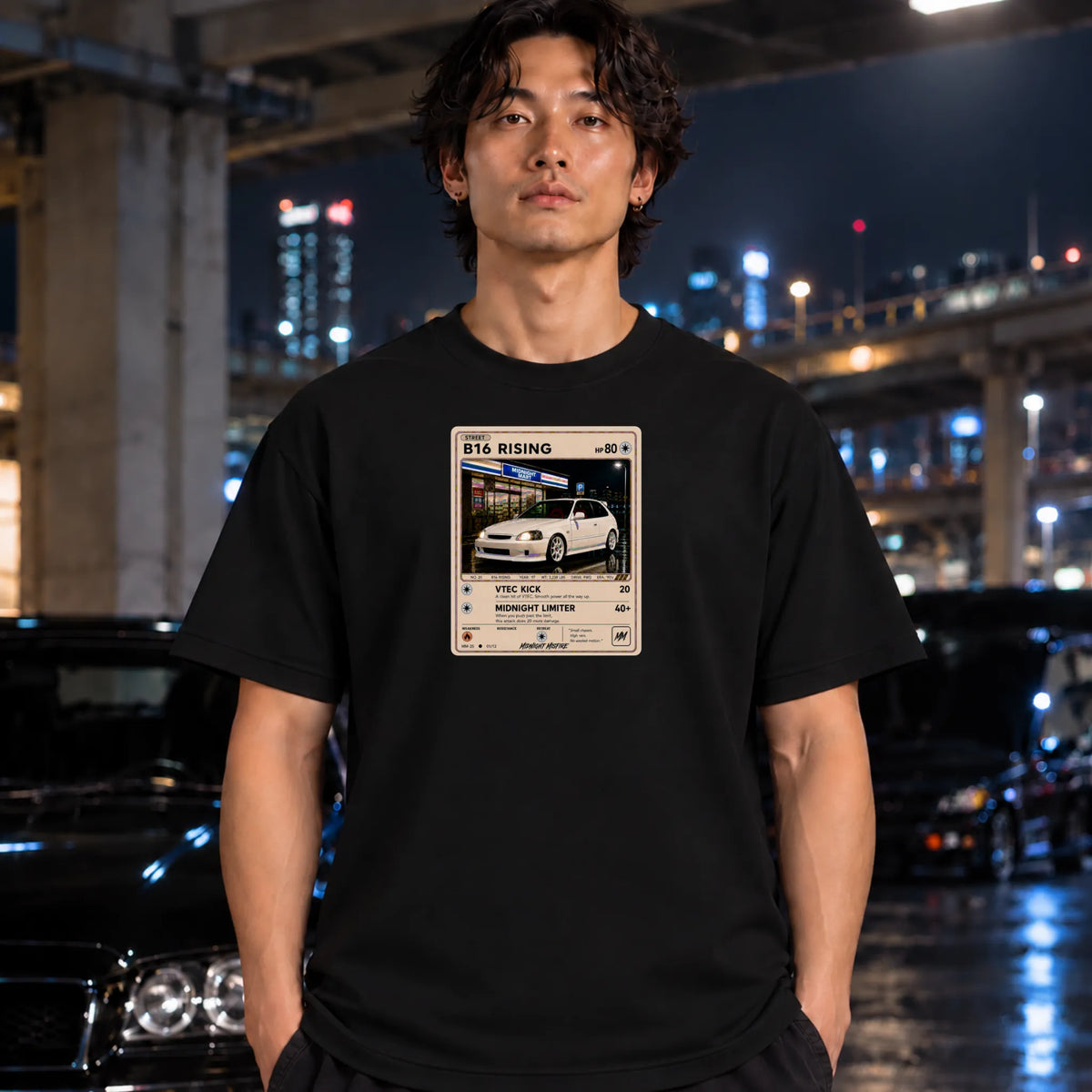 A man wearing a black t-shirt with a vintage-style ticket graphic on the chest, standing in a wet, city street at night with blurred cars and lights in the background.
