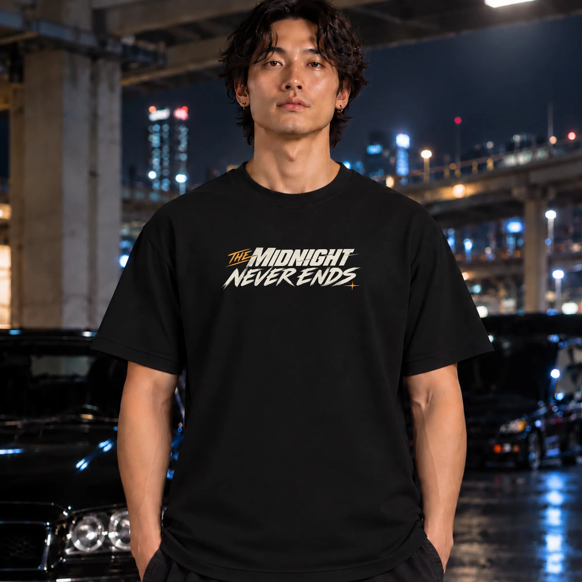 A man wearing a black t-shirt with the text 'THE MIDNIGHT NEVER ENDS' printed on the chest, standing in a wet urban night scene with cars and city lights in the background.