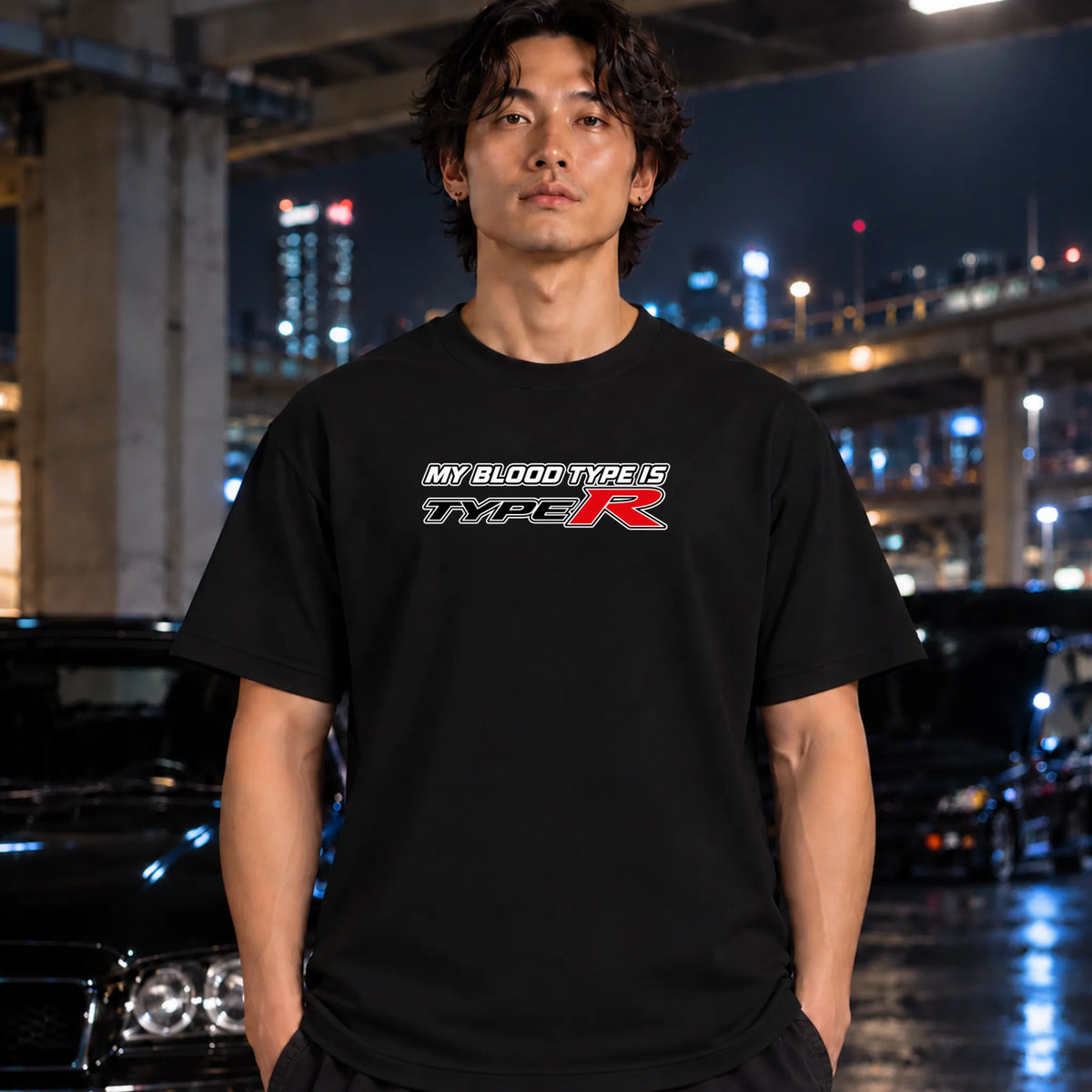 A man wearing a black t-shirt with a 'MY BLOOD TYPE IS TYPE R' graphic on the chest, standing in a wet urban night setting with cars and city lights in the background.