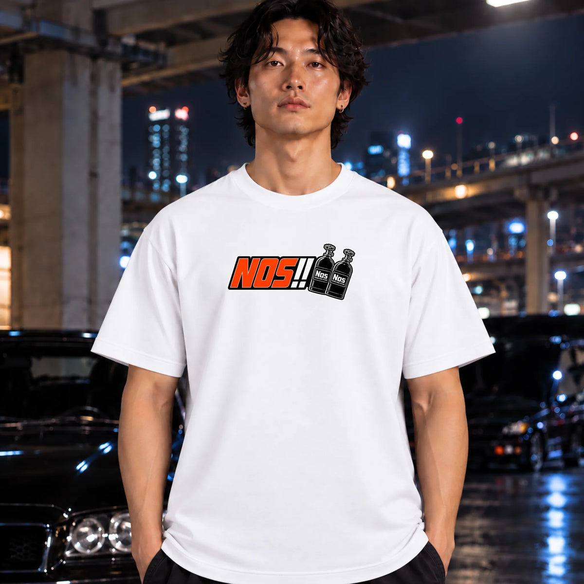 A man wearing a white short-sleeve t-shirt with a 'NOS!!' graphic on the chest, standing in front of a blurred nighttime city and bridge background with cars.