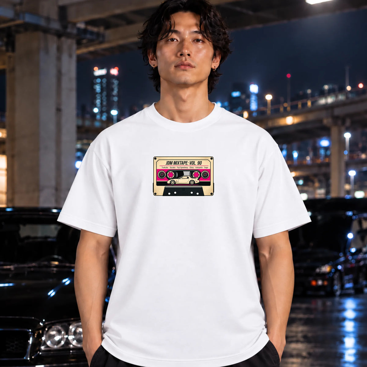 A man wearing a white graphic t-shirt with a 'JDM VOL MIXt' design, standing outdoors at night in front of a blurred city background with cars.