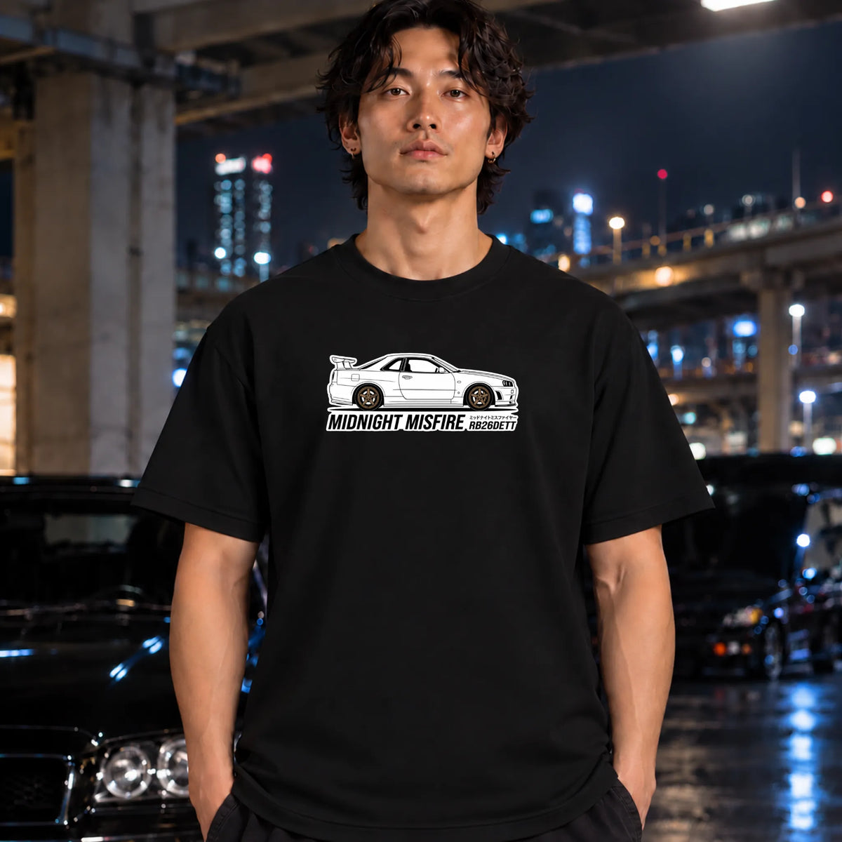 A man wearing a black t-shirt with a white graphic of a sports car and the text 'MIDNIGHT MISFIRE' stands in a city parking structure at night.