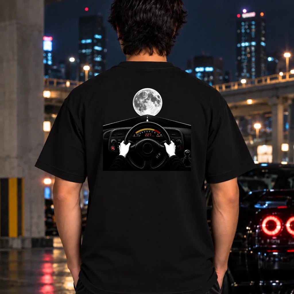 A man viewed from behind wearing a black t-shirt with a graphic print of a steering wheel, hands, and a full moon against a city night background.