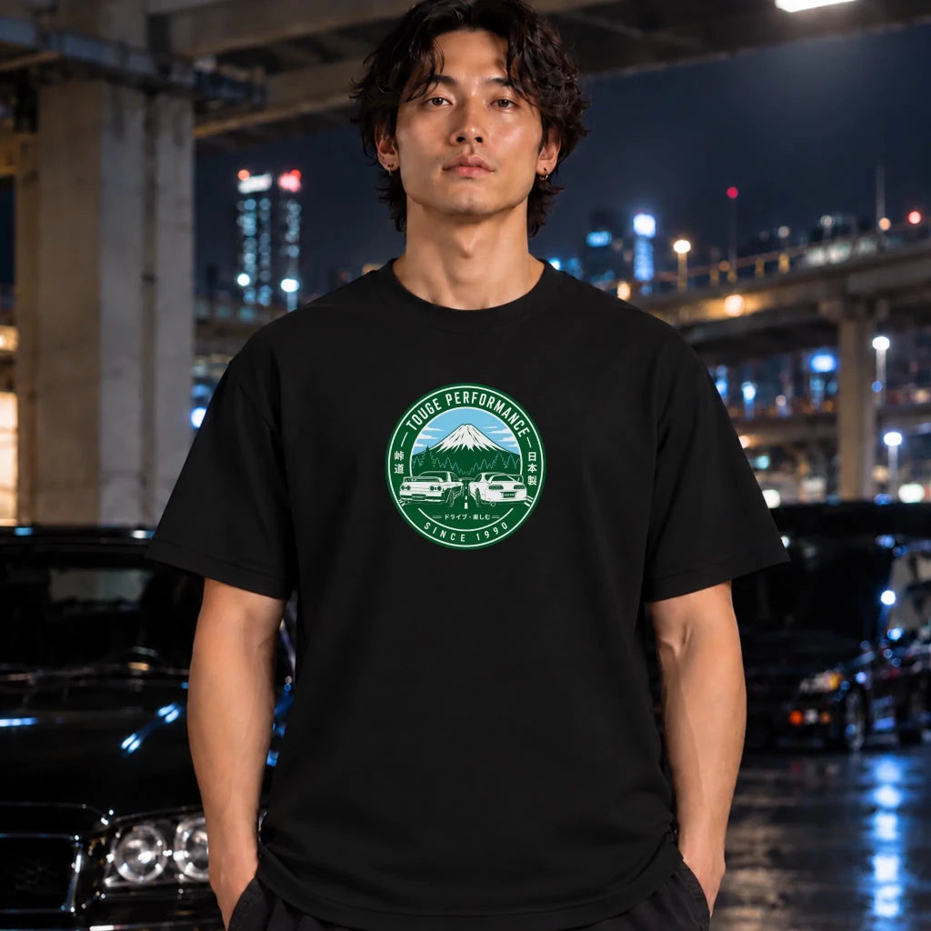 A man wearing a black t-shirt with a green circular graphic on the chest, standing in a nighttime urban setting with blurred cars and city lights in the background.
