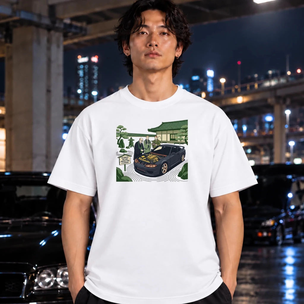 A man wearing a white graphic t-shirt with a detailed illustration of a black sports car parked in front of a Japanese temple, set against a blurred nighttime urban background with cars and city lights.