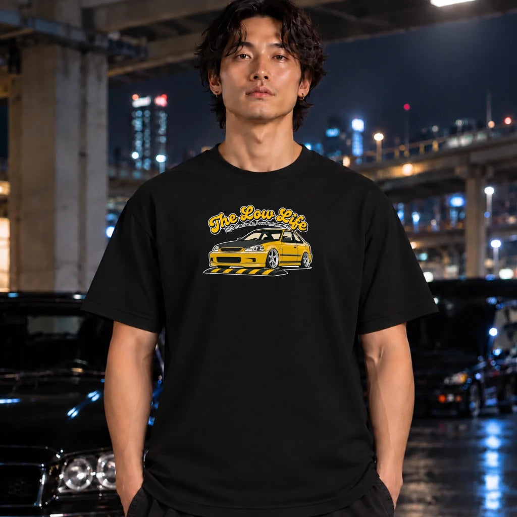A man wearing a black graphic t-shirt with a yellow and white car design and the text 'The Low Life' in a city setting at night with blurred cars and lights in the background.